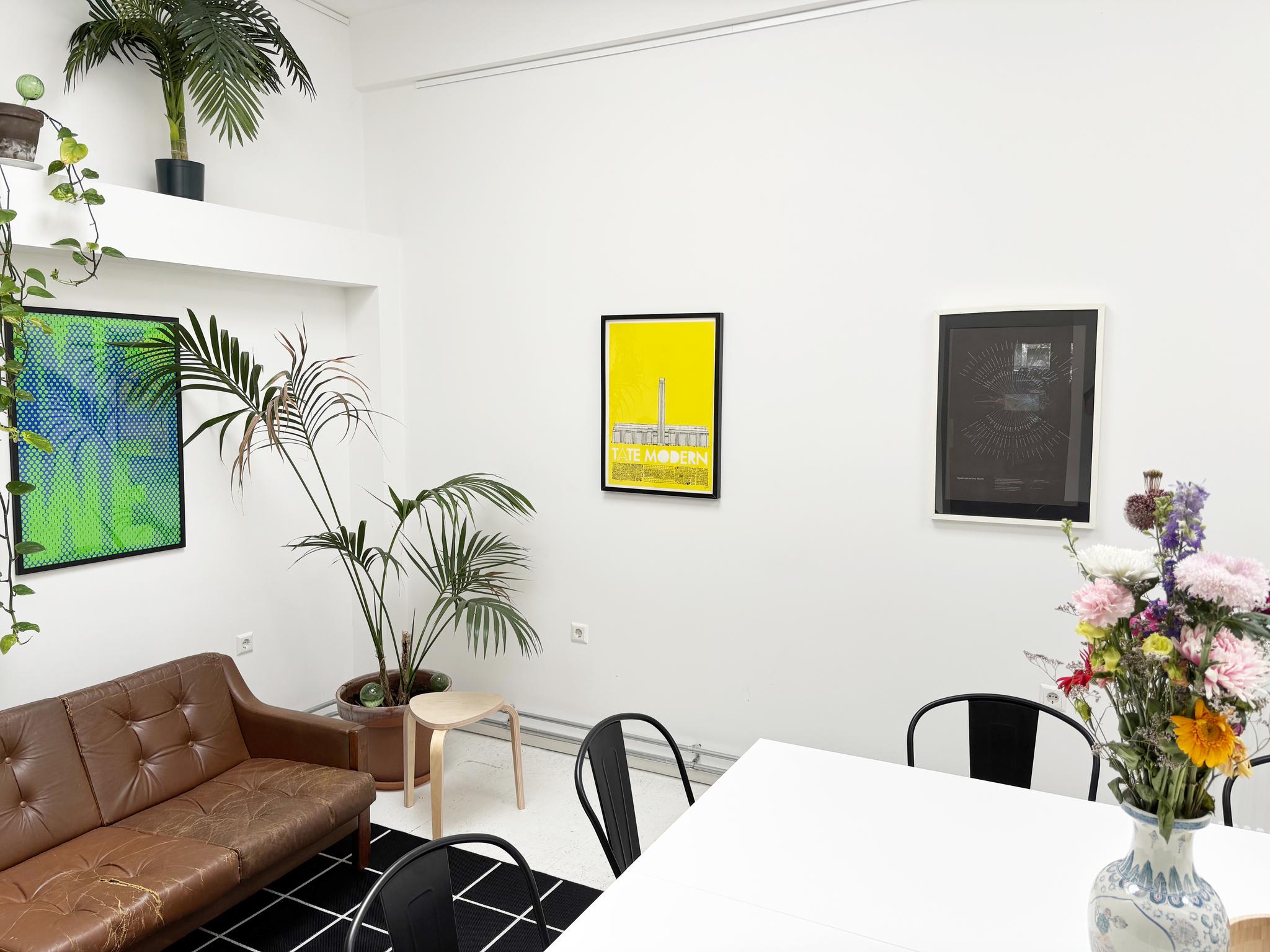 Moderne witte vergaderruimte met planten, kunst aan de muur, een bruine leren bank en een vaas met kleurrijke bloemen op tafel.